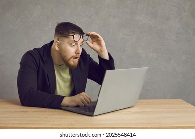 Young man is shocked while teleworking. A man in a black jacket and glasses looks in surprise at the laptop monitor with wide eyes.