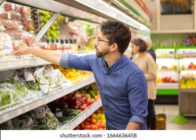 Young Man Shopping In Grocery Store. Side View Of Focused Man And Woman Holding Shopping Baskets And Choosing Fresh Fruits And Vegetables In Supermarket. Shopping Concept