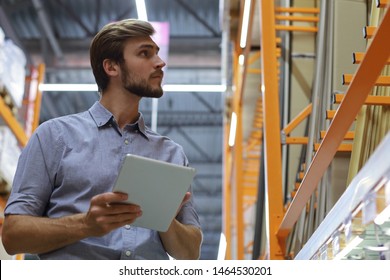 Young Man Shopping Or Working In A Hardware Warehouse Standing Checking Supplies On His Tablet.