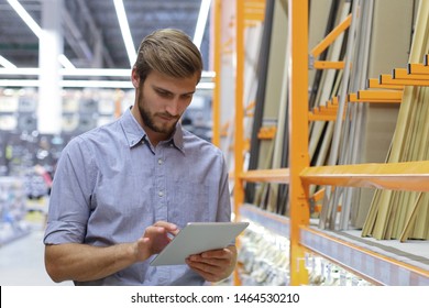 Young Man Shopping Or Working In A Hardware Warehouse Standing Checking Supplies On His Tablet.