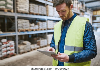 Young man shopping or working in a hardware warehouse standing checking supplies on his tablet with an absorbed expression