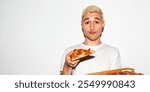 Young man with short blond hair holding a slice of pizza. Casual pose with pizza box. Enjoying pizza, wearing a white t-shirt, against a plain background. Blonde man eating a pizza from a box.