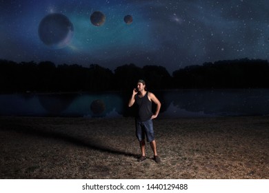 Young Man In Shorts And Cap, At Night Talking On A Mobile Phone On The Shore Of A Reservoir Against The Background Of An Abstract Night Sky