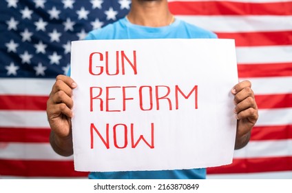 Young Man Showing Gun Reform Now Sign Board In Front Of US American Flag - Concept Of Protest Against Gun Violence And Activism.