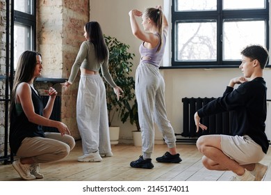 Young man showing new vogue dance movements to one of active teenage girls sitting in front of him and asking questions