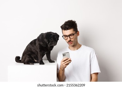 Young Man Showing Photos On Smartphone To Cute Dog, Pet Owner And Pug Staring At Mobile Phone, White Background