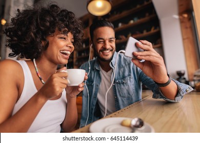 Young Man Showing Something On Cellphone To His Girlfriend At Cafe. Happy Young Couple Sitting At Coffee Shop Having Video Chat On Mobile Phone.