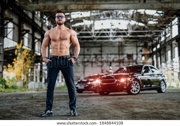 The young\
man shows beautiful body. Guy with nacked torso poses at abandoned\
warehouse with luxury car\
background.