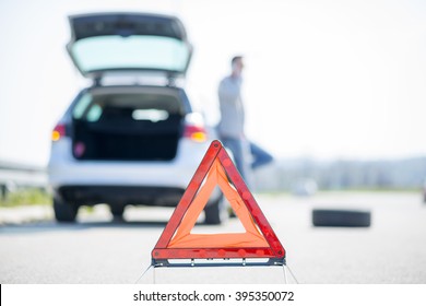 A young man with a silver car that broke down on the road.He has set up a warning triangle.He is waiting for the technician to arrive.