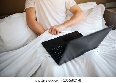 Young man sit in bed early morning. He hold hand under white blanket and masturbating. Laptop on his legs. Watching video.