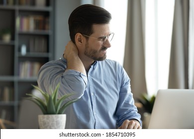 Young man sit at workplace desk touch neck feeling ache, massaging nape to relief painful feelings. Poor sitting posture during laptop usage, sedentary lifestyle, chronic disorder fibromyalgia concept - Shutterstock ID 1854710395