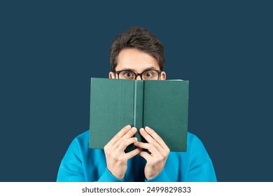 A young man sits against a solid dark blue background, holding an open book in front of his face. He wears glasses and a bright blue sweater, peering over the top of the book - Powered by Shutterstock