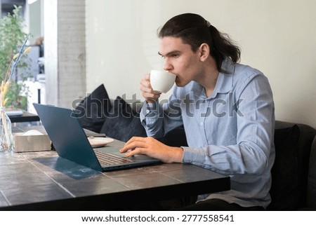A young man sits at a cafe table focused on his laptop while sipping coffee. The scene takes place in a cozy cafe with plants and soft lighting on a weekday morning.