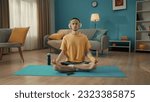 A young man sits cross legged in a lotus position with his eyes closed on a sports mat next to a smartphone. A man in a tracksuit and white wireless headphones is doing yoga at home.