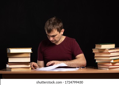 A Young Man Sits At A Desk Surrounded By Books And Sorting Through Papers. Student Is Preparing For The Exam. Freelancer At Work. Black Background. Isolate.