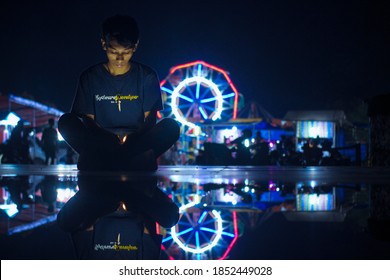 A Young Man Sits Near A Puddle Of Water And Plays With His Cell Phone In The Background Of The City Night Lights, 24 May 2019, Kendari City, Southeast Sulawesi, Indonesia