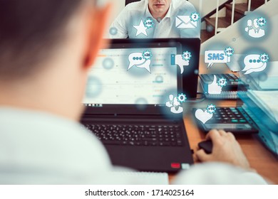 A young man sits in a new modern office at a table in a laptop and holds a mobile phone in his hands. Icons of social networks fly out of the gadget. Social network technology concepts. - Shutterstock ID 1714025164