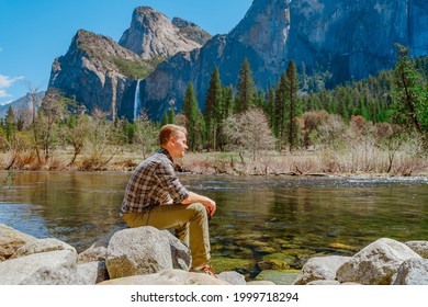 A young man sits on the bank of a river and enjoys the view of a mountain waterfall in Yosemite National Park
