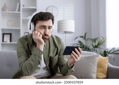 Young man sits on couch looking bored while using phone, wearing headphones. Concept of boredom, technology, relaxation, and indoor lifestyle. Captures everyday moments of using modern devices. - Powered by Shutterstock