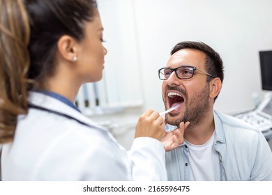 A Young Man Sits On An Exam Table Across From His Doctor. The Doctor Reaches Forward With A Tongue Depressor As The Man Looks Up And Sticks Out His Tongue.