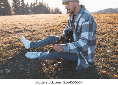 Young man sits on a grassy field in the warm autumn sunlight, smiling as he plays the ukulele. Surrounded by golden tones and distant trees, the moment reflects joy, creativity, and a deep connection. - Powered by Shutterstock