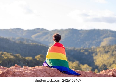 A young man sits on a mountain peak, wrapped in a rainbow flag, symbolizing LGBT pride month, with a backdrop of  mountains - Powered by Shutterstock