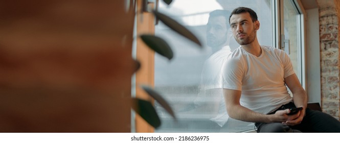 A young man sits on a windowsill and looks at the city on a rainy day. A handsome, pumped up brunette guy sits thoughtfully by a large window at his home on a cloudy day. - Powered by Shutterstock
