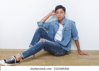 A young man sits on a wooden background wearing a blue shirts and torn jeans with sneakers


