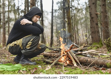 A young man sits in the woods by a burning fire with his arms folded across his chest. The tourist is dressed in a black jacket and green hiking pants. - Powered by Shutterstock