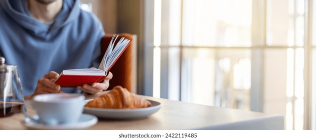 A young man is sitting alone in a cozy cafe, engrossed in a captivating book. The aroma of freshly brewed coffee wafts through the air as he enjoys a warm croissant on the table. - Powered by Shutterstock
