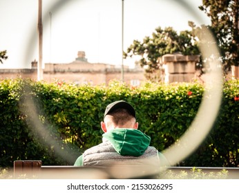 A young man sitting alone in the park as seen through the fence