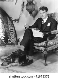 Young Man Sitting In An Armchair Next To A Fire Place With His Dog By His Feet