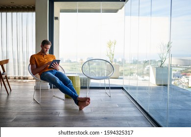 Young Man Sitting Back In A Chair In His Living Room At Home Browsing Online With A Digital Tablet