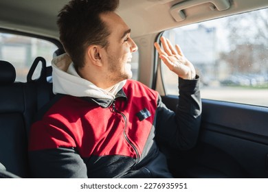 A young man sitting in the back seat of car while using his phone and waving through the window