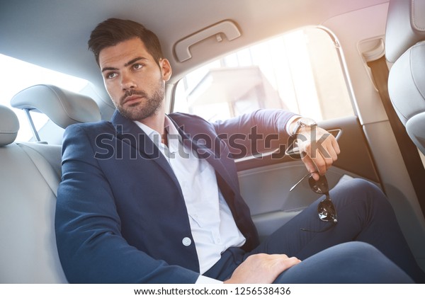 A young man is sitting\
in the backseat of cars and looks out the window. In his hands,\
sunglasses