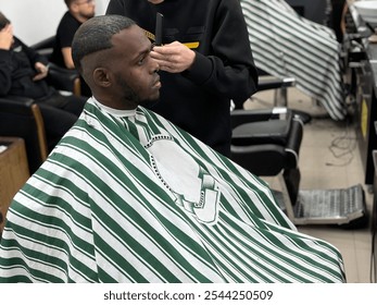 Young man sitting in a barbershop chair, getting a stylish haircut. Focused barber with clippers and scissors, creating a modern look. Perfect image for concepts of grooming, style, and personal care. - Powered by Shutterstock