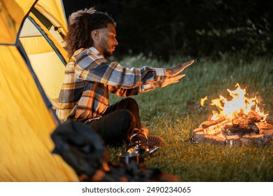 Young man sitting by a campfire, warming his hands outside a tent in the forest at night. - Powered by Shutterstock