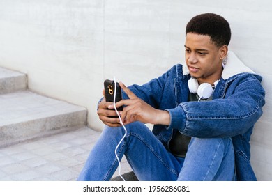 Young Man Sitting Connected To The Internet With His Smart Phone. Confident Guy Sending A Message To A Friend. Young Afro Boy With Headphones On The Outside.