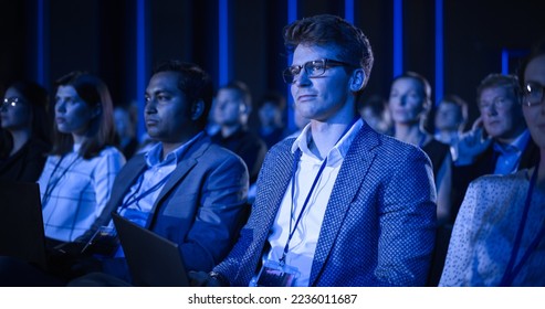 Young Man Sitting in a Crowded Audience at an IT Conference. Delegate Using Laptop Computer to Take Notes. Software Developer Watching Presentation About New High Tech Products and Cloud Solutions. - Powered by Shutterstock