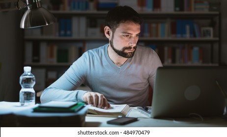 Young Man Sitting At Desk, Using A Laptop, Working Late At Night, He Is Studying A Book And Using A Laptop