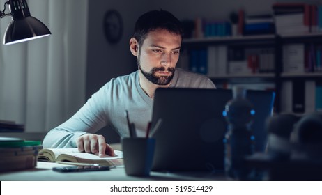 Young Man Sitting At Desk, Using A Laptop, Working Late At Night, He Is Studying A Book And Using A Laptop