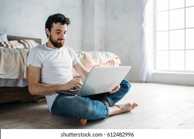 Young Man Sitting Down On The Floor Near The Bed With Folded Legs. Nice Cute Guy Is Working Or Watching Interesting Video On The Laptop. He Is Smiling