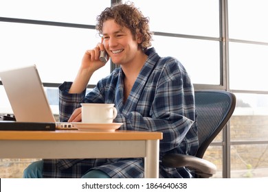 Young man sitting at his desk in his home office and talking on the telephone while wearing a dressing gown and smiling