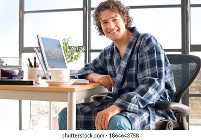 Young Man Sitting At His Desk In His Home Office And Leaning On The Table While Wearing A Dressing Gown And Smiling