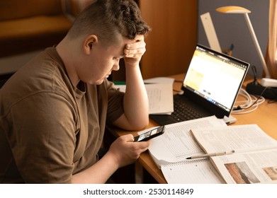 A young man is sitting at his desk at home in front of a laptop, looking at his mobile phone. Various papers are spread out in front of him. - Powered by Shutterstock