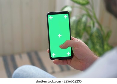 Young man sitting at home holding smartphone green mock-up screen in hand. Male person using chroma key mobile phone. Vertical mode. Touching, swiping display, tapping, surfing Internet social media - Powered by Shutterstock