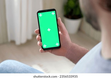 Young man sitting at home holding smartphone green mock-up screen in hand. Male person using chroma key mobile phone. Vertical mode. Touching, swiping display, tapping, surfing Internet social media - Powered by Shutterstock