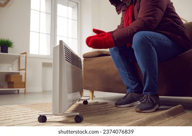 Young man is sitting at home on the sofa in warm jacket, knitted mittens and scarf near the heater, freezing from the cold. There is no heat in the house, there are problems with heating.