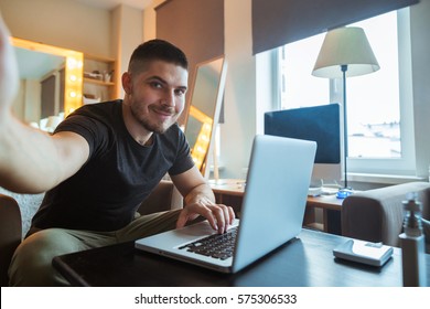 Young Man Sitting At A Laptop A Freelancer In The Office Does Selfie. Home Office.