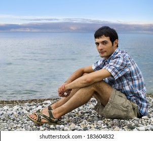 young man sitting looking in camera on pebble beach during sunset
